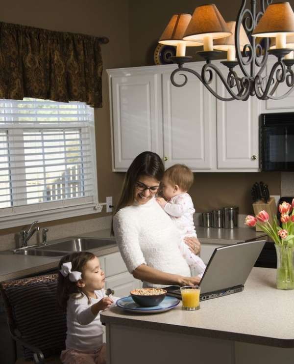 Mother holding baby and typing on laptop computer with girl eating breakfast in kitchen.